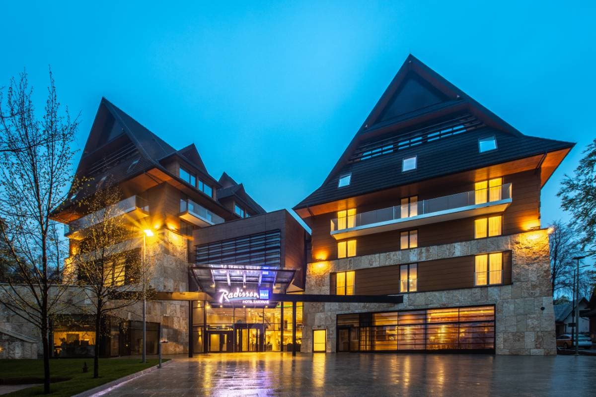 Radisson Residences Zakopane — elewacja budynku o zmierzchu z oświetlonymi oknami, drewnianą fasadą i znakiem Radisson Blu